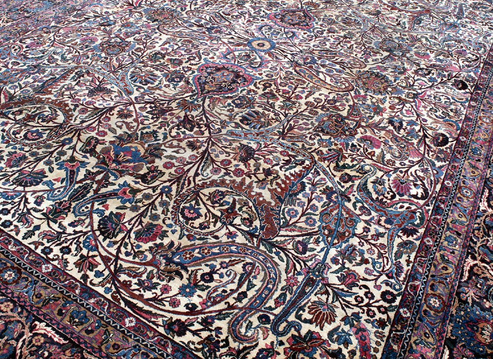 A large Kirman wool carpet featuring intricate floral scrollwork, paisley boteh motifs, and a central medallion in cream, magenta, and navy blue tones. View V