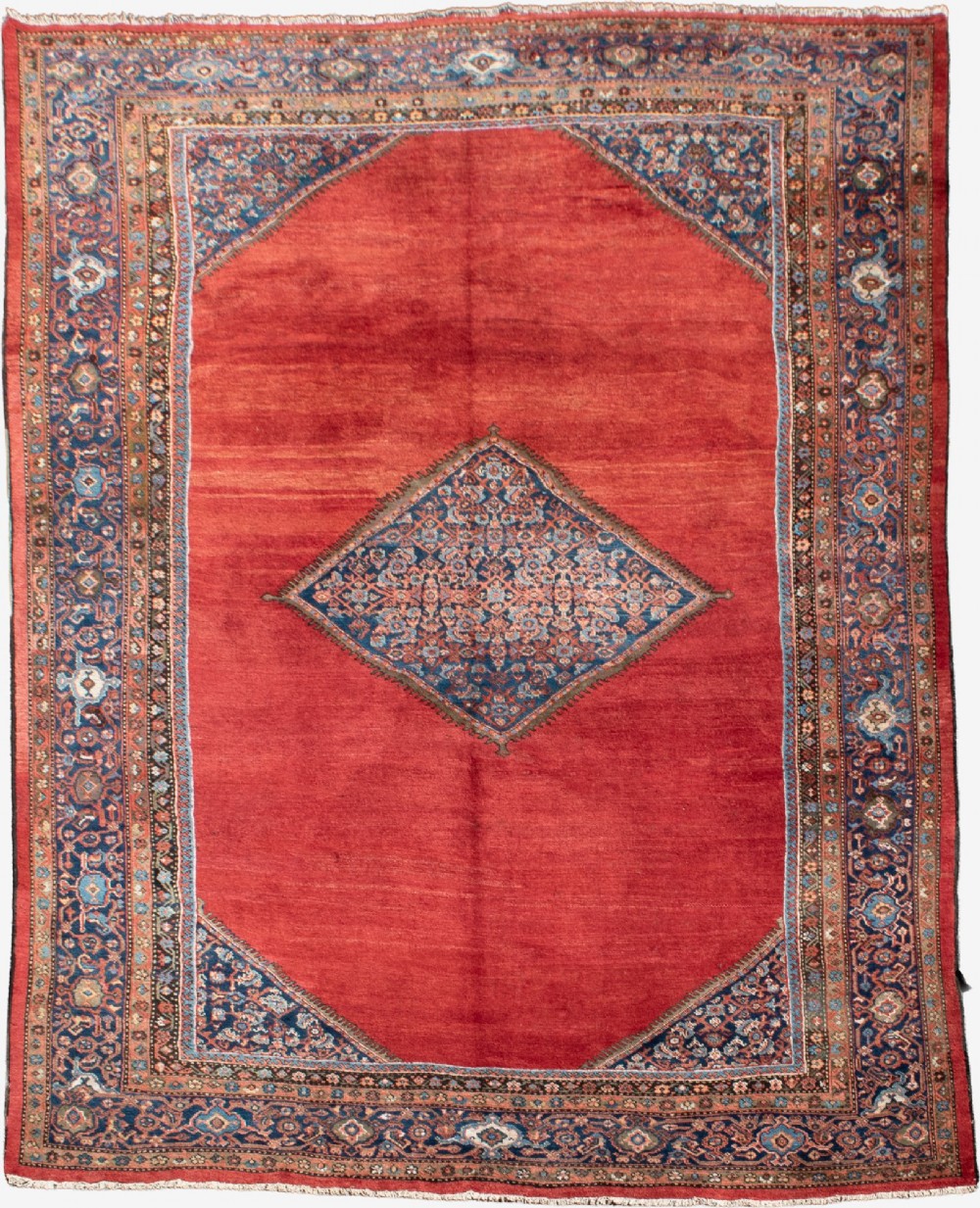 A large hand-knotted antique Ferahan wool carpet featuring a terracotta red field, a central diamond-shaped blue medallion, and detailed ornamental borders.