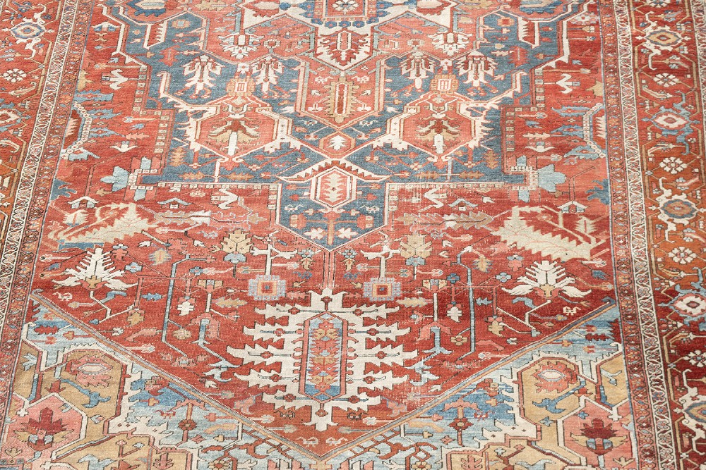 An antique Persian Heriz wool carpet featuring a bold geometric indigo medallion set against a terracotta red field with stepped spandrels and ornate borders. View III