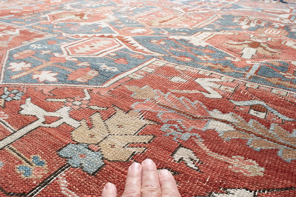An antique Persian Heriz wool carpet featuring a bold geometric indigo medallion set against a terracotta red field with stepped spandrels and ornate borders. View X