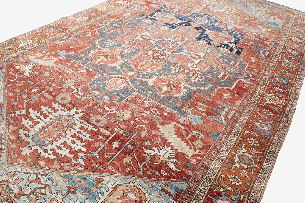 An antique Persian Heriz wool carpet featuring a bold geometric indigo medallion set against a terracotta red field with stepped spandrels and ornate borders. View XII