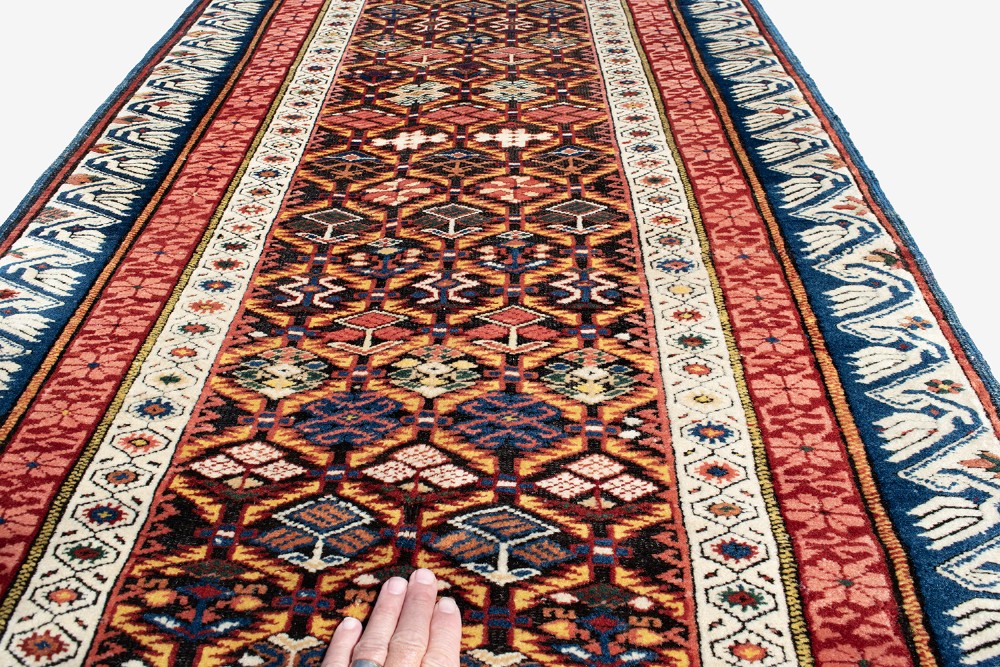 A long hand-knotted Caucasian Seychor wool rug runner featuring a dark central field with repeating geometric diamond motifs, flanked by red decorative inner borders and a blue wave pattern outer border. View III