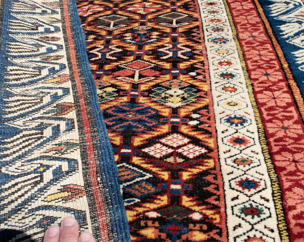 A long hand-knotted Caucasian Seychor wool rug runner featuring a dark central field with repeating geometric diamond motifs, flanked by red decorative inner borders and a blue wave pattern outer border. View IV