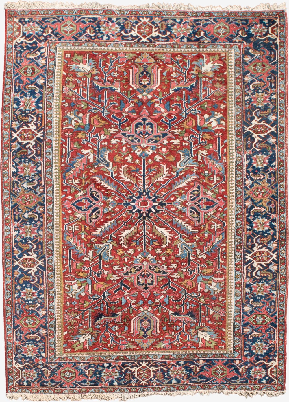 A large hand-knotted antique Persian Heriz wool carpet featuring a bold central medallion surrounded by geometric floral motifs in red, blue, green, and ivory tones.