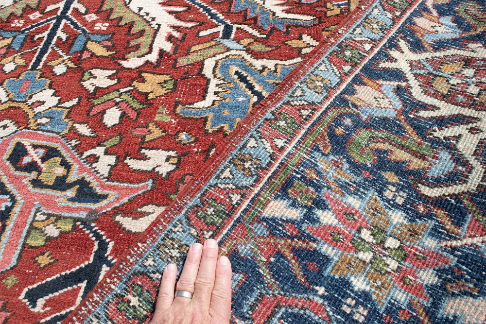 A large hand-knotted antique Persian Heriz wool carpet featuring a bold central medallion surrounded by geometric floral motifs in red, blue, green, and ivory tones. View VI