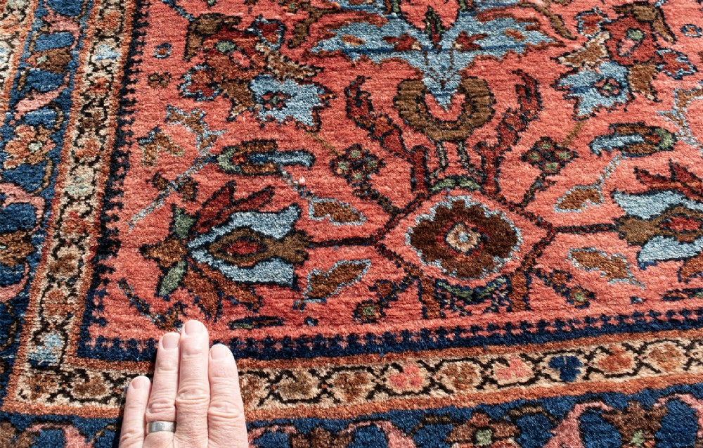 A small rectangular hand-knotted wool rug featuring a coral-red central field decorated with blue and brown floral motifs surrounded by a dark blue border. View III