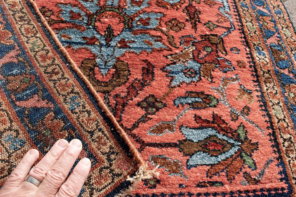 A small rectangular hand-knotted wool rug featuring a coral-red central field decorated with blue and brown floral motifs surrounded by a dark blue border. View IV