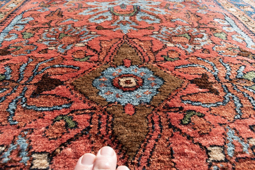 A small rectangular hand-knotted wool rug featuring a coral-red central field decorated with blue and brown floral motifs surrounded by a dark blue border. View V