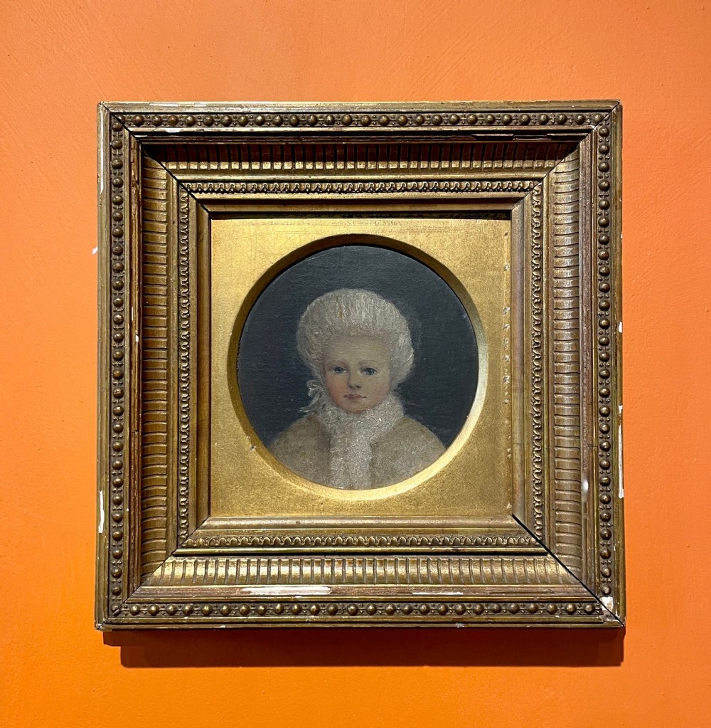 A Victorian oil portrait painting of a young girl wearing a white fur-trimmed cap inside a circular aperture framed by a square gilded moulding.