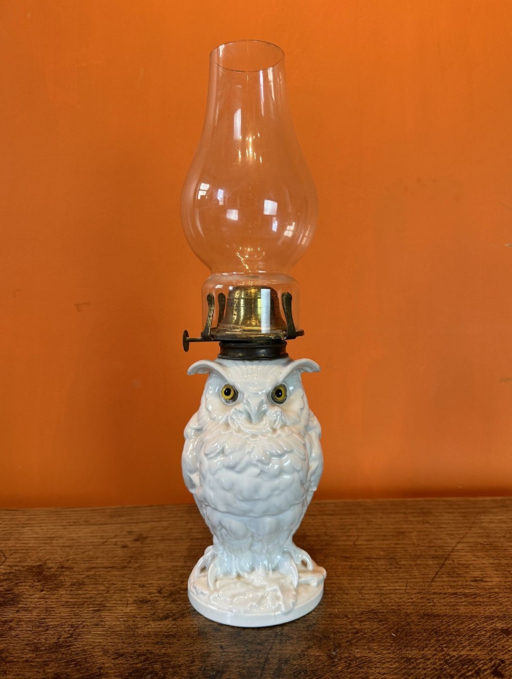A Victorian white glazed porcelain oil lamp shaped like an owl with glass eyes, a brass burner, and a clear glass chimney.