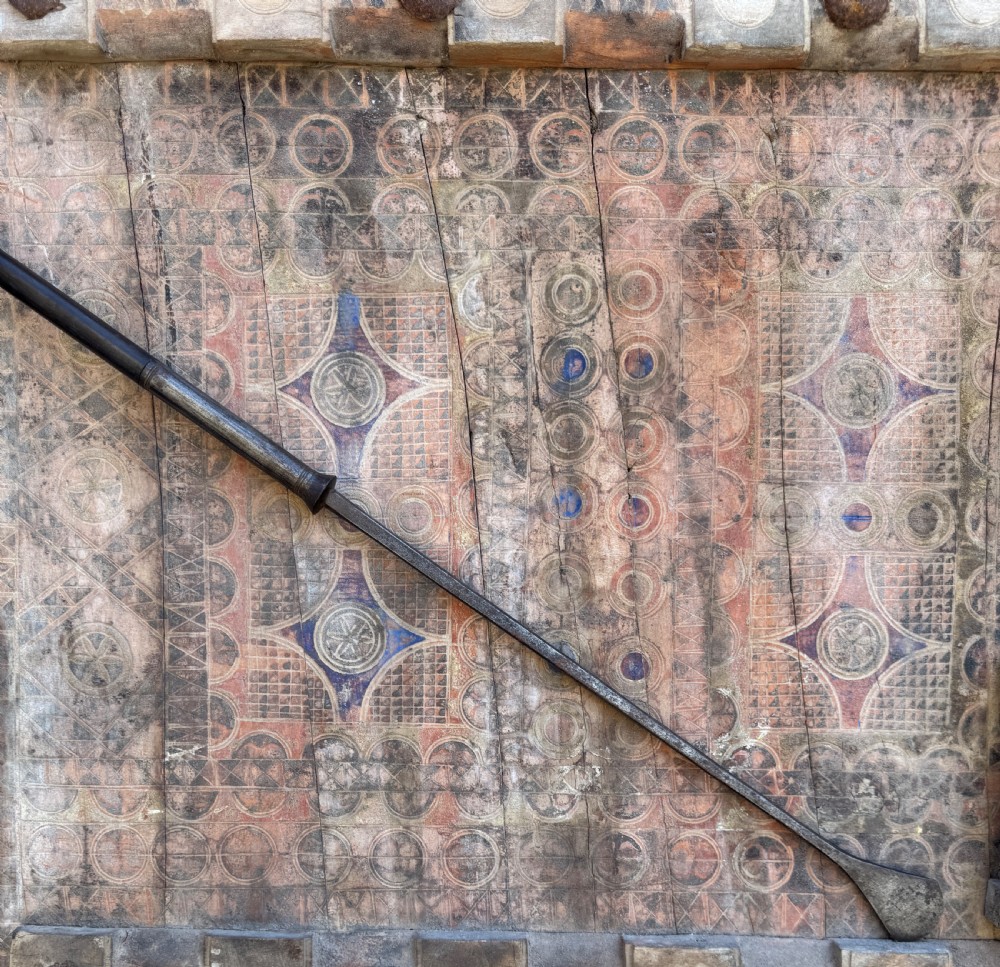 A hand-forged dark steel spearhead with a defined central ridge attached to a slender wooden shaft. View IV