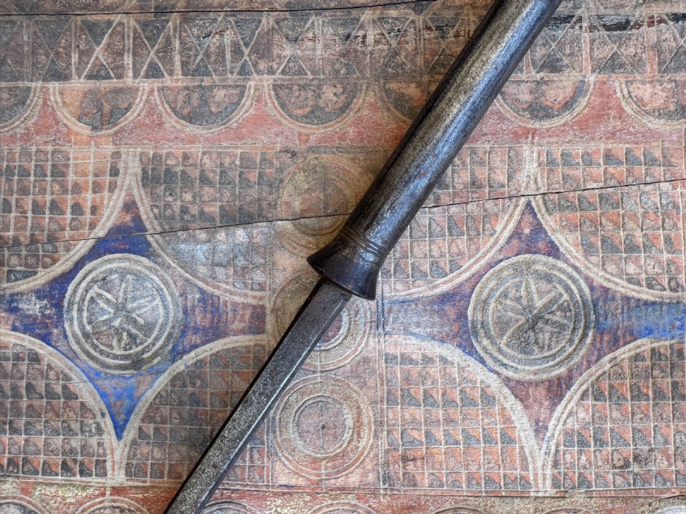 A hand-forged dark steel spearhead with a defined central ridge attached to a slender wooden shaft. View V