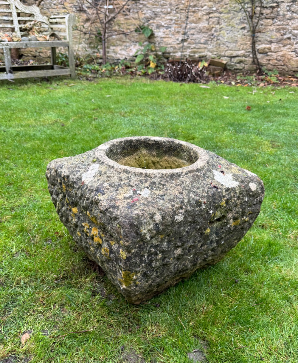 A square carved weathered stone planter with a circular central recess and rusticated hand-chiselled exterior surfaces displaying natural moss growth.