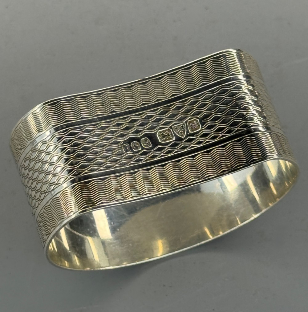 An oval antique sterling silver napkin ring featuring detailed engine-turned wavy patterns and a central circular cartouche engraved with initials. View III