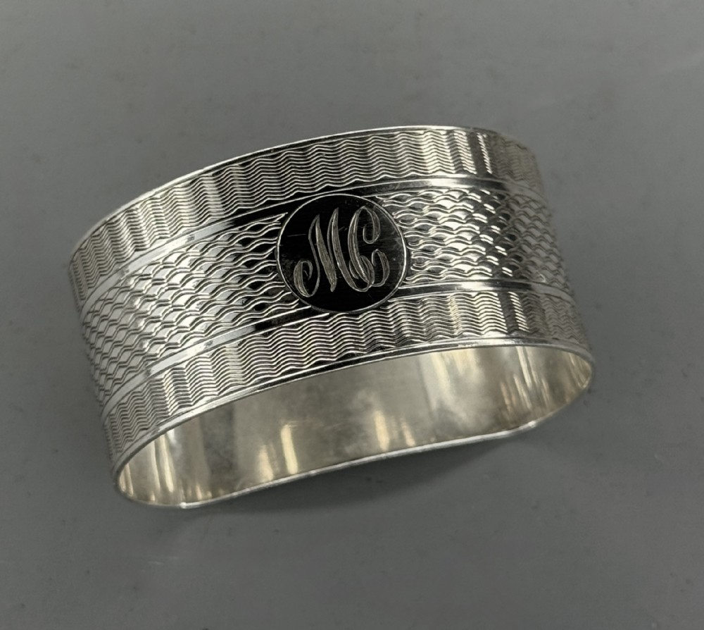 An oval antique sterling silver napkin ring featuring detailed engine-turned wavy patterns and a central circular cartouche engraved with initials.