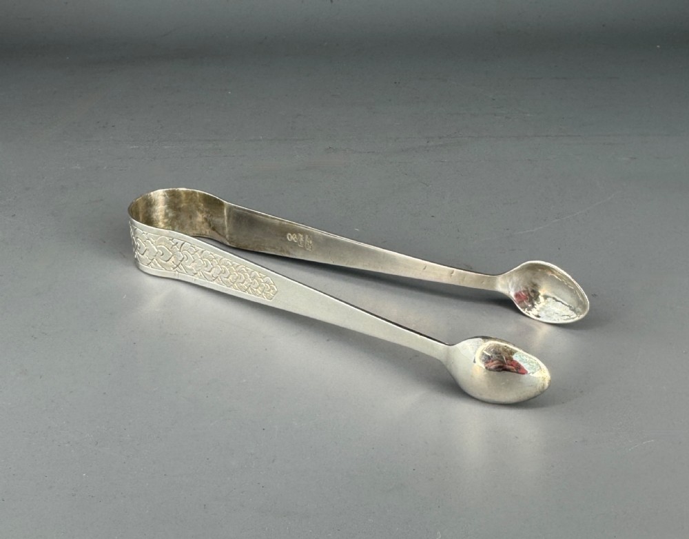 A pair of silver sugar tongs featuring textured engraved decoration near the arch and spoon-shaped bowls.