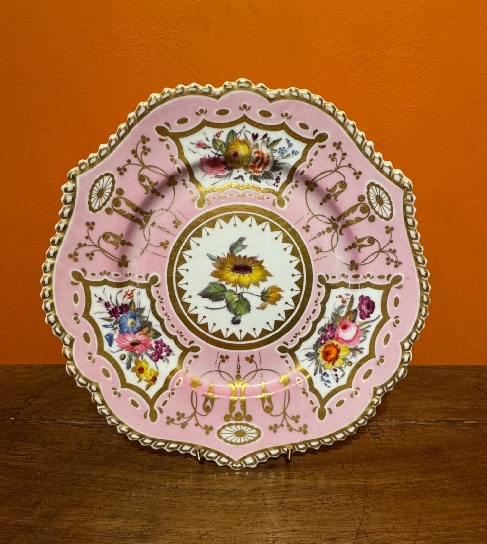 An early 19th-century circular porcelain cabinet plate featuring a pink ground, scalloped gold rim, hand-painted central sunflower, and surrounding floral cartouches with gilt detailing.