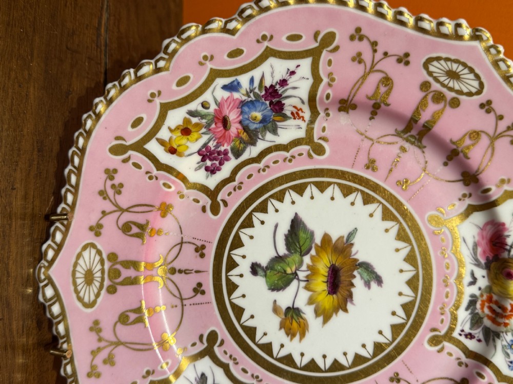 An early 19th-century circular porcelain cabinet plate featuring a pink ground, scalloped gold rim, hand-painted central sunflower, and surrounding floral cartouches with gilt detailing. View IV