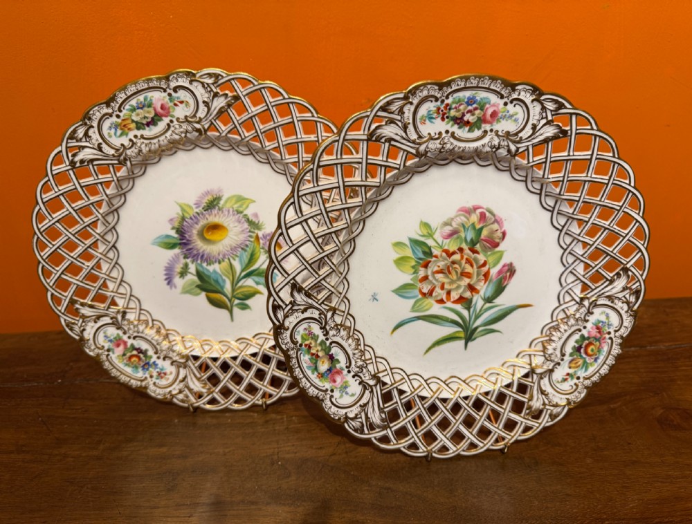Pair of circular white porcelain plates with reticulated basketweave borders, gilded scrollwork, and hand-painted central botanical sprays.