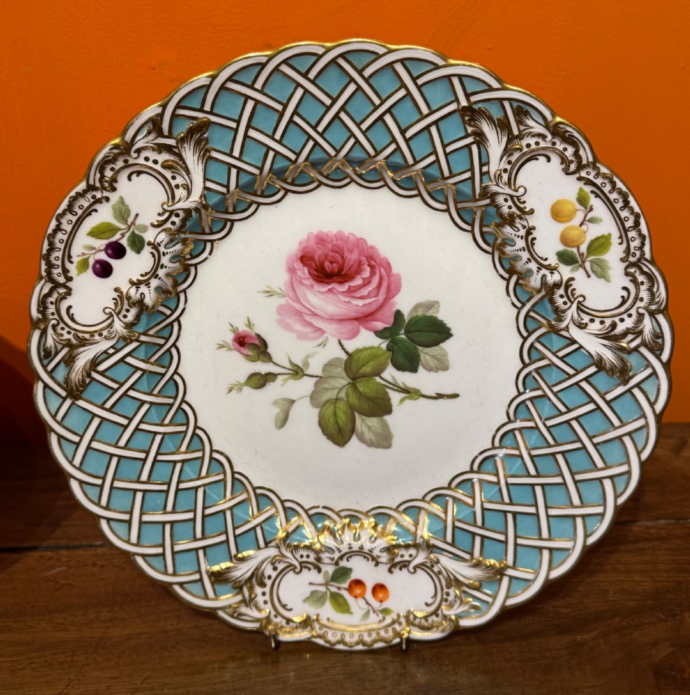 A circular Victorian Minton porcelain plate featuring a central hand-painted pink rose surrounded by a turquoise basketweave border and gilded scroll cartouches displaying fruit motifs.