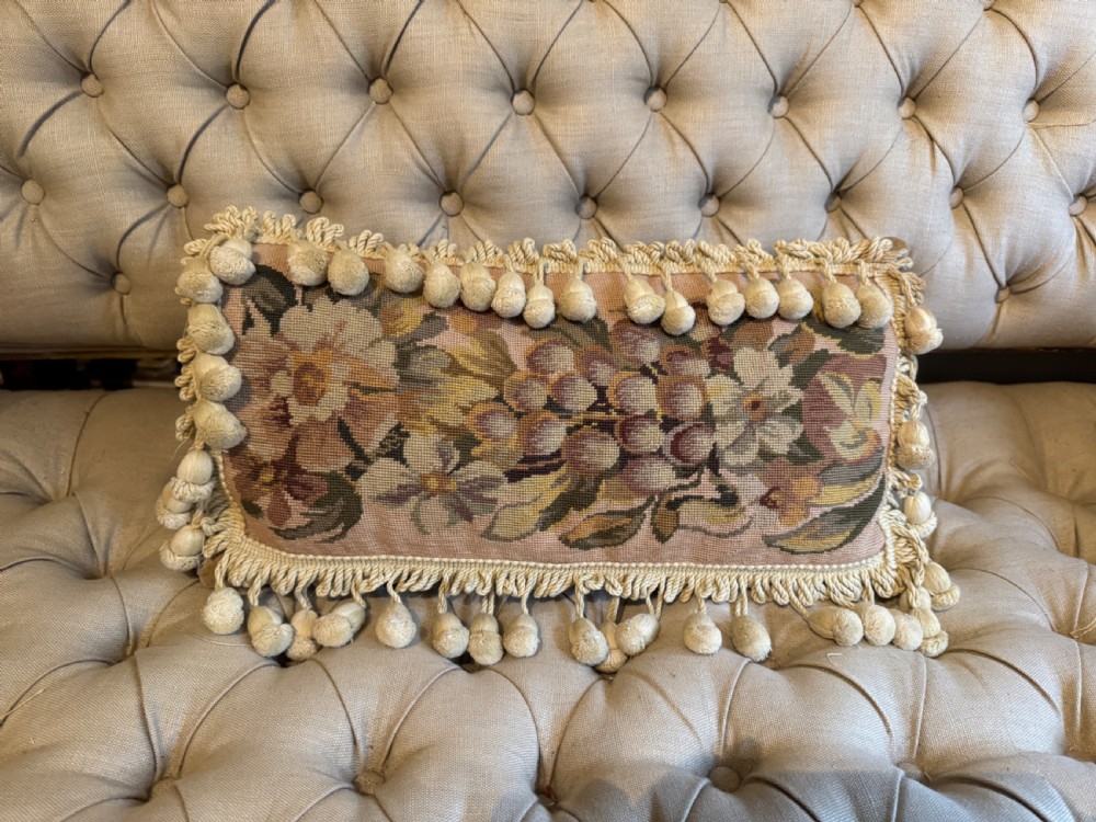 A rectangular antique French tapestry cushion with floral needlepoint detail in beige and pink tones, trimmed with a fringe and pom-pom border.