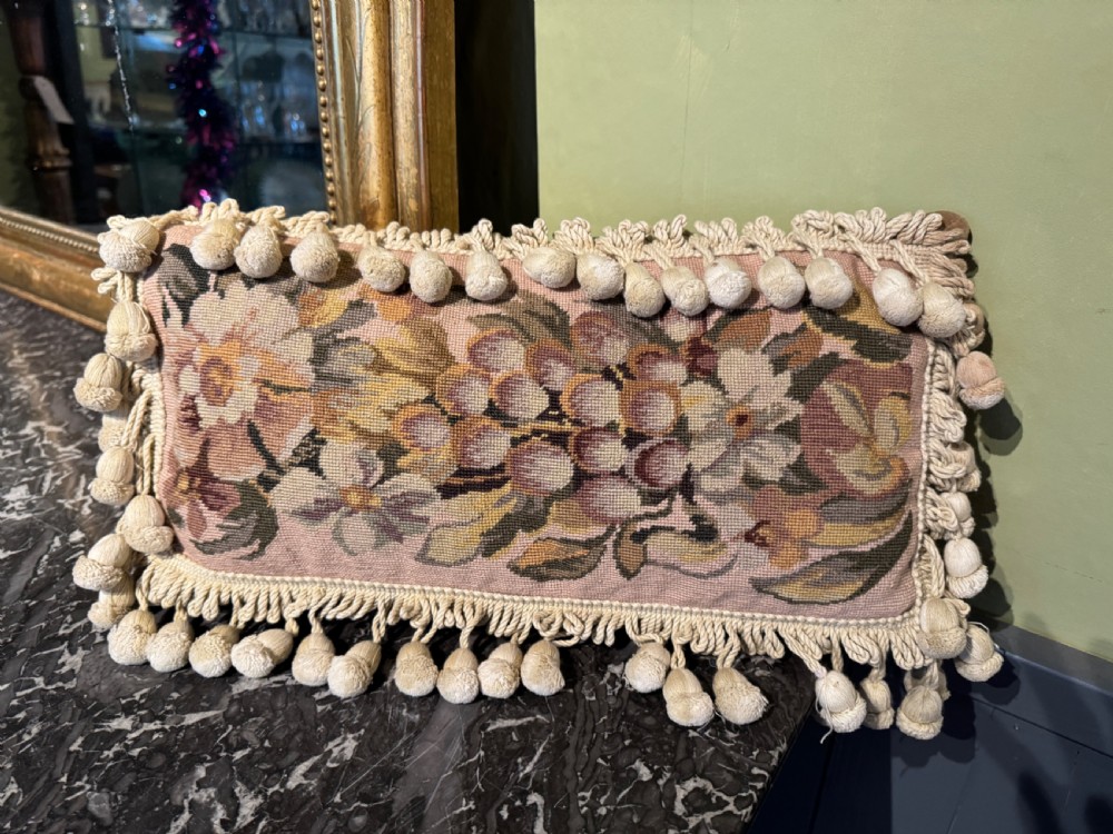 A rectangular antique French tapestry cushion with floral needlepoint detail in beige and pink tones, trimmed with a fringe and pom-pom border. View V
