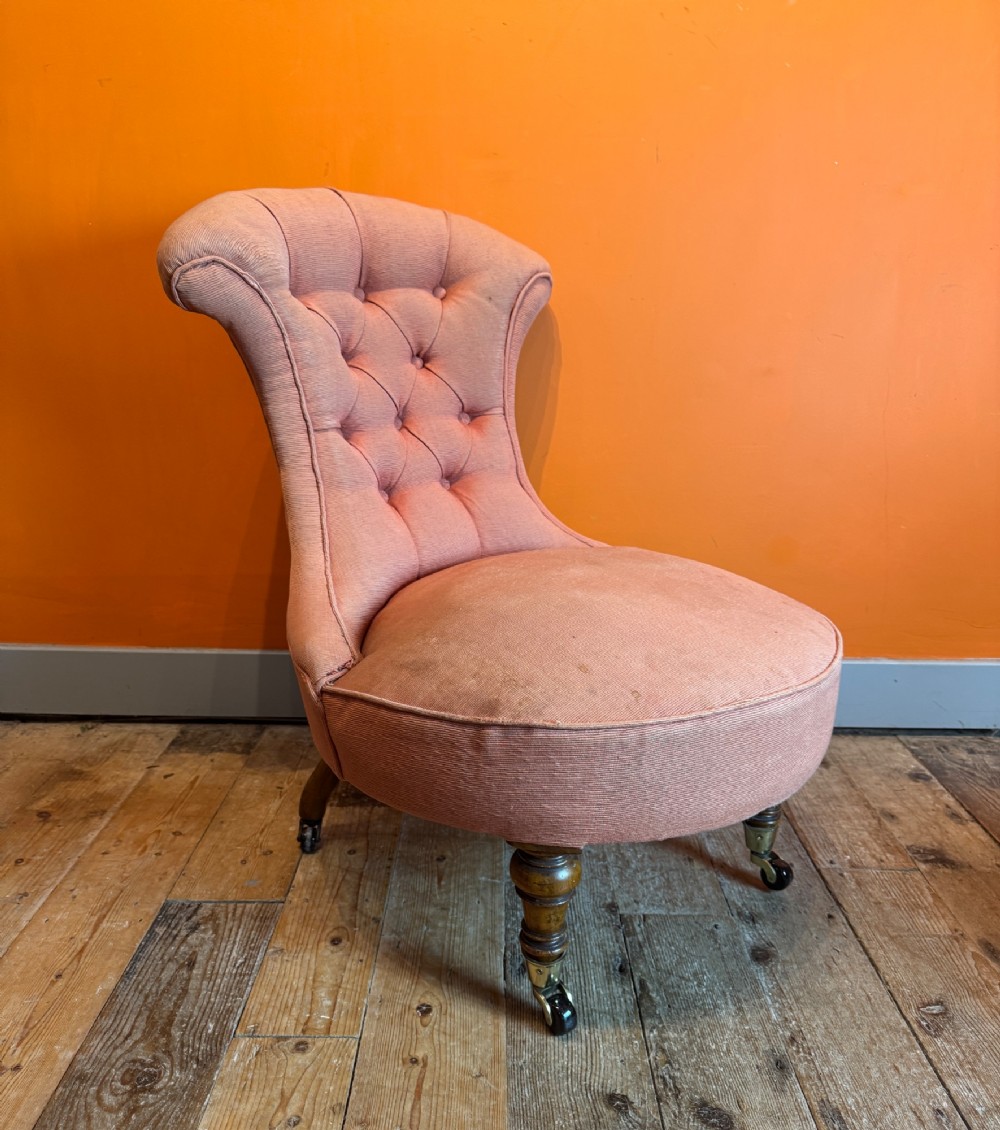 An antique Victorian button-back slipper chair with low salmon pink upholstery, turned wooden front legs, and brass castors.