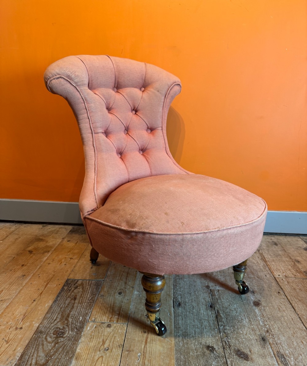 An antique Victorian button-back slipper chair with low salmon pink upholstery, turned wooden front legs, and brass castors. View II