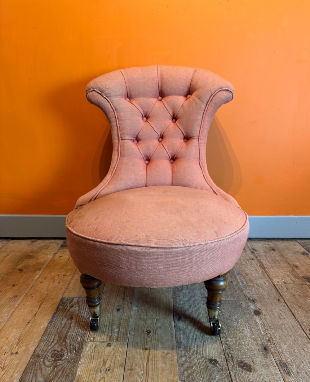 An antique Victorian button-back slipper chair with low salmon pink upholstery, turned wooden front legs, and brass castors. View III