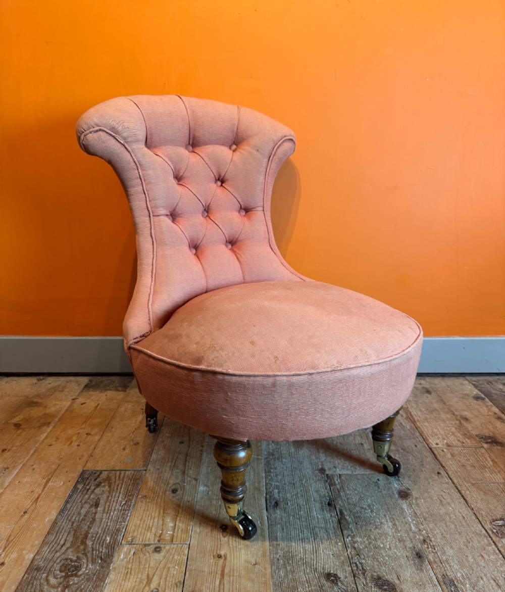 An antique Victorian button-back slipper chair with low salmon pink upholstery, turned wooden front legs, and brass castors. View IV