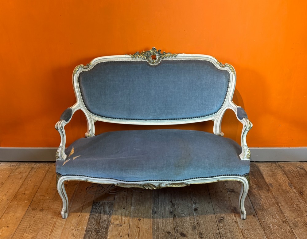 An antique Louis XV style two-seat settee featuring a cream painted and carved wooden frame with gilt accents, cabriole legs, and blue-grey fabric upholstery.