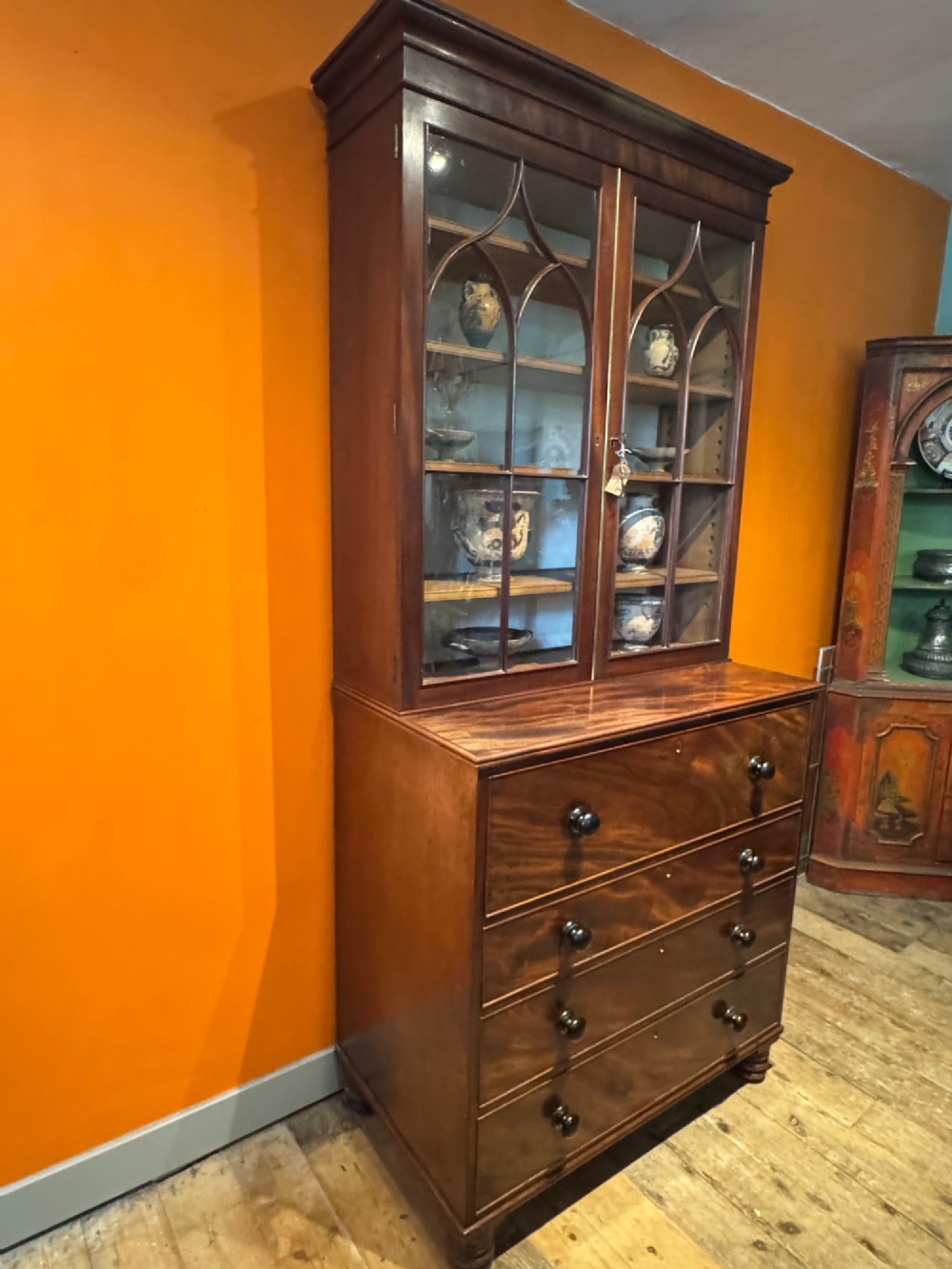 An antique Regency mahogany secretaire bookcase featuring upper Gothic astragal glazed doors over a lower chest of drawers on turned feet. View V