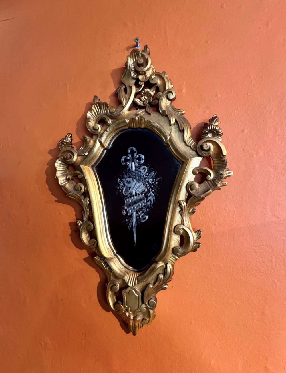 An Italian Rococo carved giltwood wall mirror with an ornate C-scroll frame and an engraved mirror plate displaying a musical instrument motif.