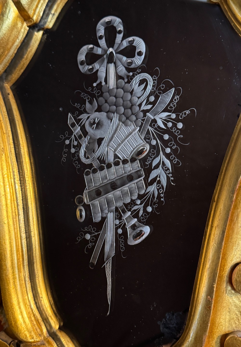 An Italian Rococo carved giltwood wall mirror with an ornate C-scroll frame and an engraved mirror plate displaying a musical instrument motif. View V