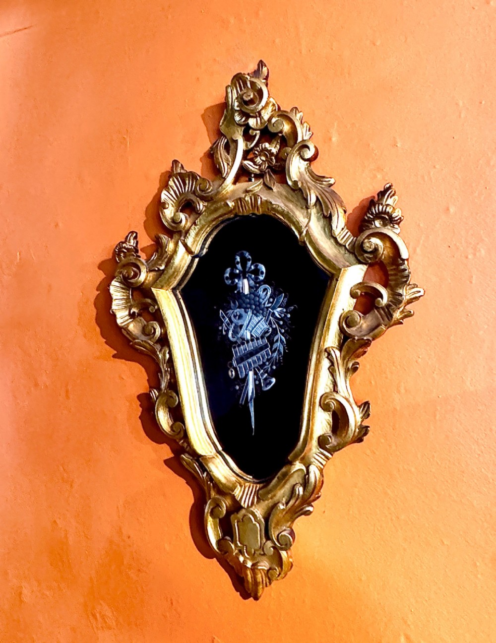 An Italian Rococo carved giltwood wall mirror with an ornate C-scroll frame and an engraved mirror plate displaying a musical instrument motif. View VI