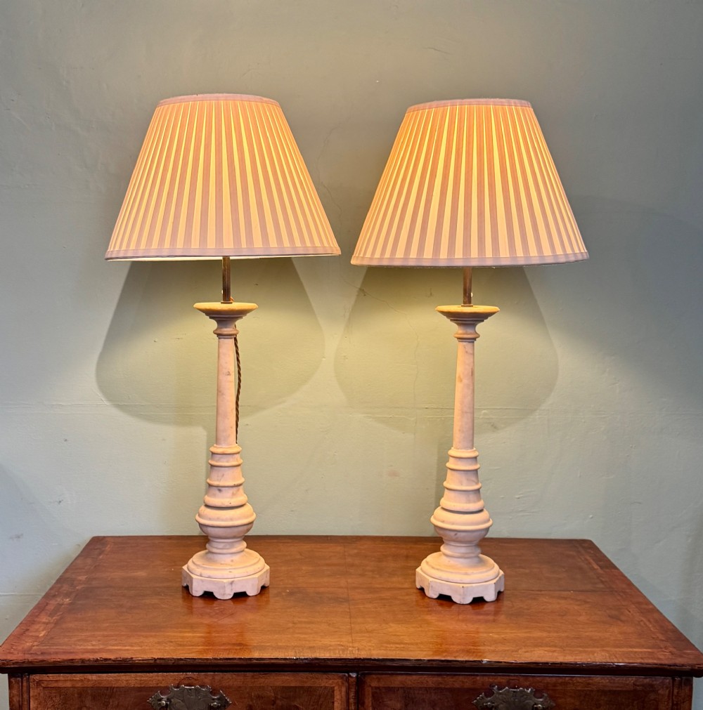 A pair of turned off-white marble table lamps featuring cylindrical baluster shafts, stepped circular bases, and cream pleated fabric shades.