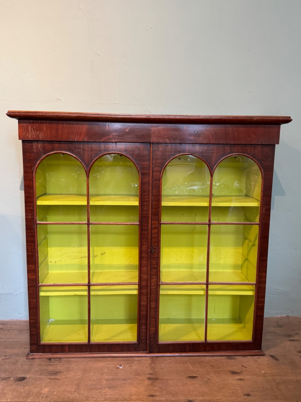 A Victorian dark mahogany two-door cabinet featuring glass doors with arched glazing bars and a bright yellow painted interior with shelves. View II