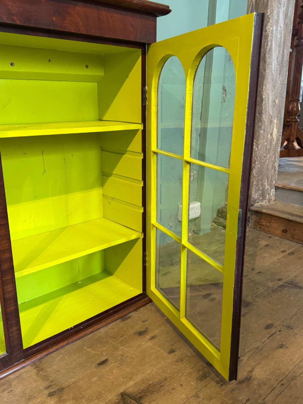 A Victorian dark mahogany two-door cabinet featuring glass doors with arched glazing bars and a bright yellow painted interior with shelves. View VI