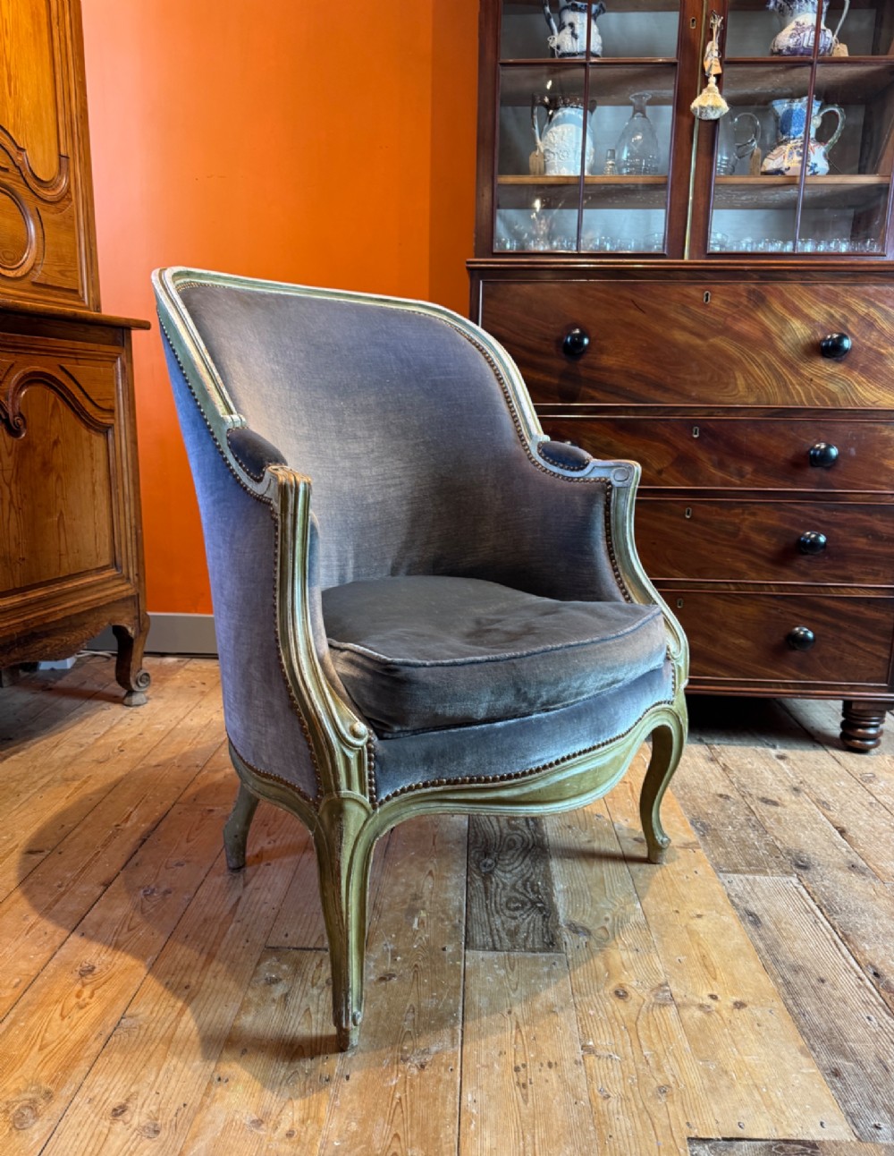 An antique Louis XV style tub bergere armchair upholstered in slate grey velvet with a carved distressed green wooden frame and cabriole legs.