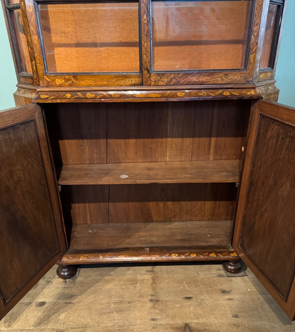 An antique Dutch walnut display cabinet featuring intricate floral marquetry inlays, glazed upper doors, and a double cupboard lower section supported by bun feet. View X