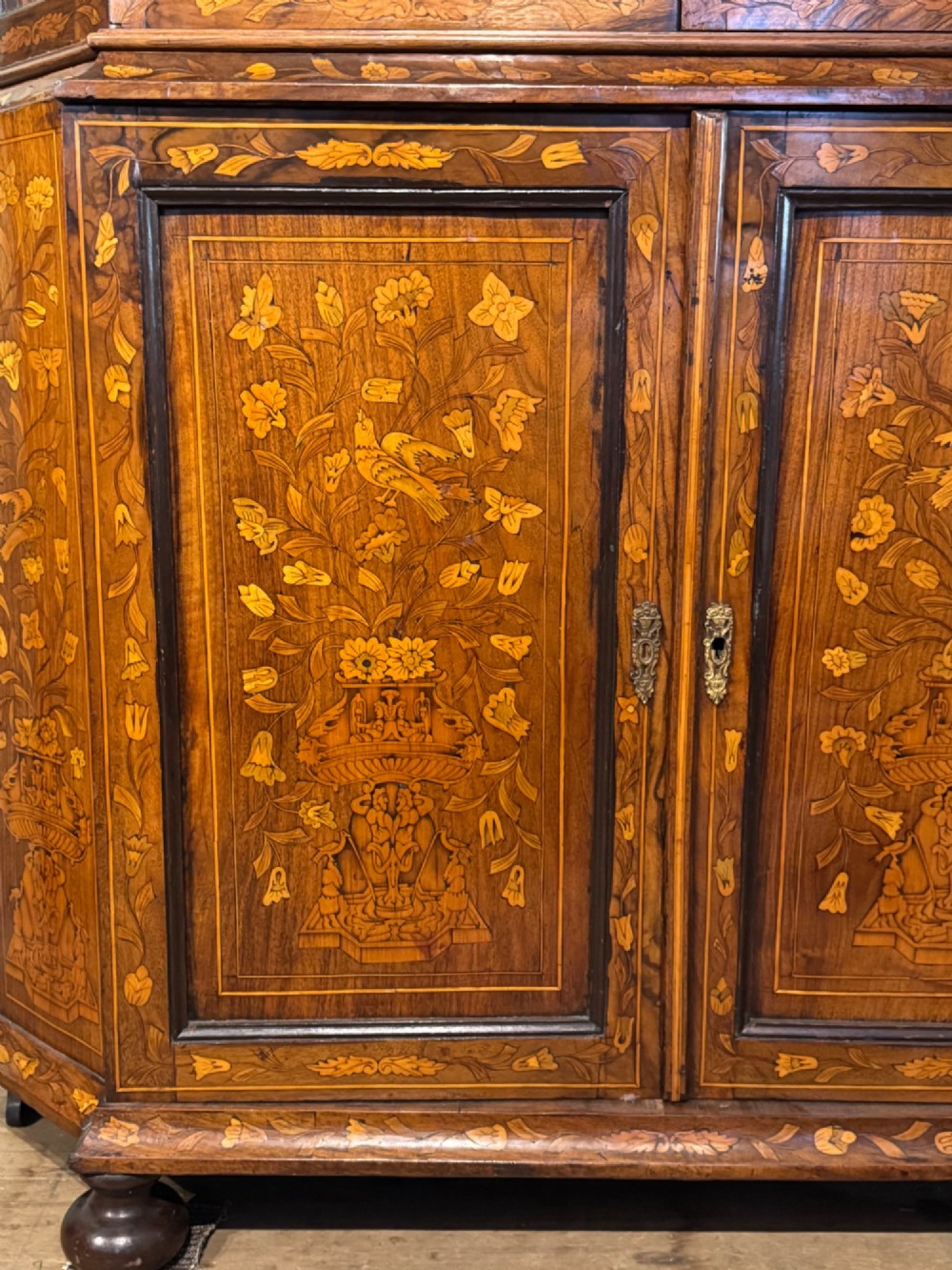 An antique Dutch walnut display cabinet featuring intricate floral marquetry inlays, glazed upper doors, and a double cupboard lower section supported by bun feet. View XII