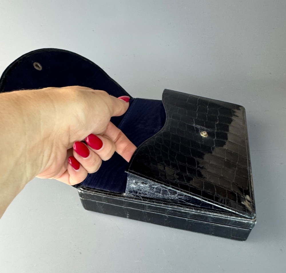 A black crocodile leather rectangular vanity case featuring a curved front flap pocket, top handle, and brass clasp. View V