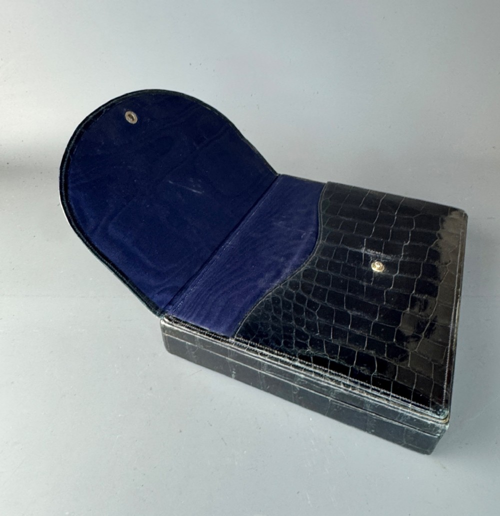 A black crocodile leather rectangular vanity case featuring a curved front flap pocket, top handle, and brass clasp. View VI