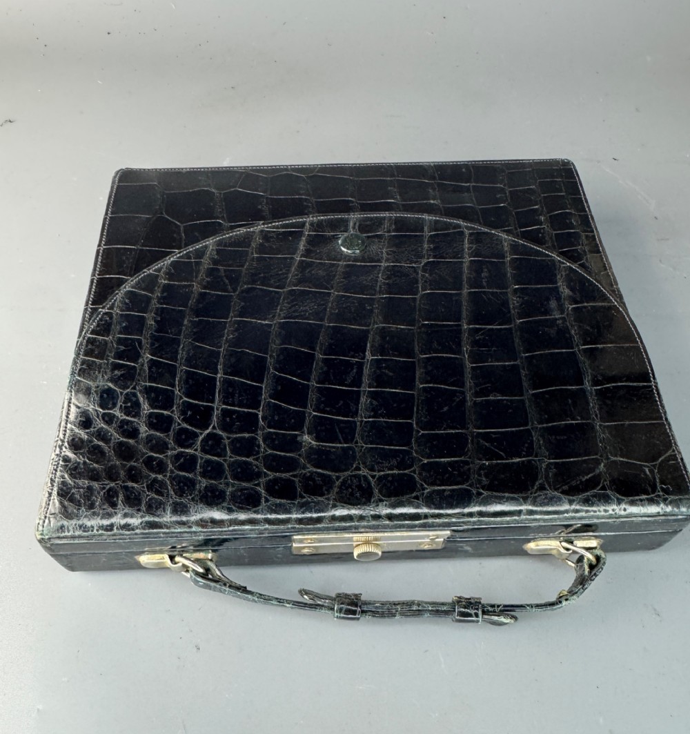 A black crocodile leather rectangular vanity case featuring a curved front flap pocket, top handle, and brass clasp. View VIII