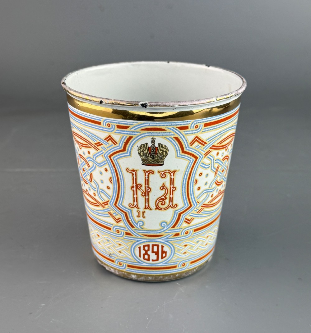 A white enamelled steel beaker decorated with red, blue, and gold geometric patterns, featuring the crowned cypher of Tsar Nicholas II and the date 1896.