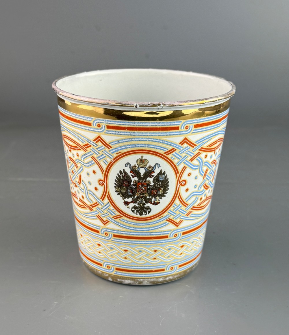 A white enamelled steel beaker decorated with red, blue, and gold geometric patterns, featuring the crowned cypher of Tsar Nicholas II and the date 1896. View II
