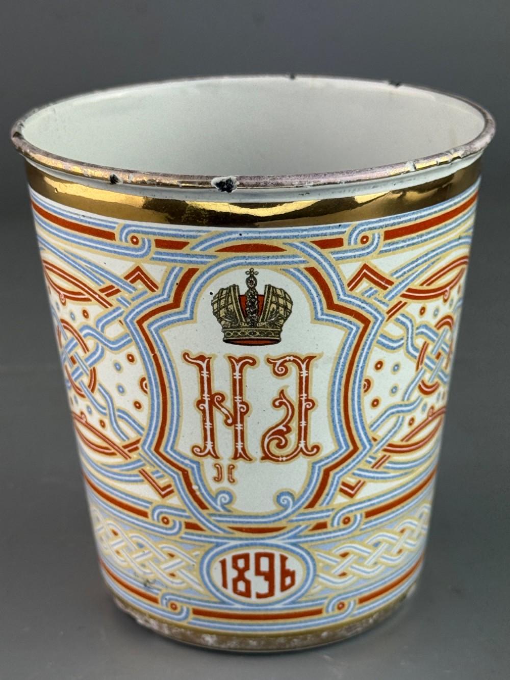 A white enamelled steel beaker decorated with red, blue, and gold geometric patterns, featuring the crowned cypher of Tsar Nicholas II and the date 1896. View III