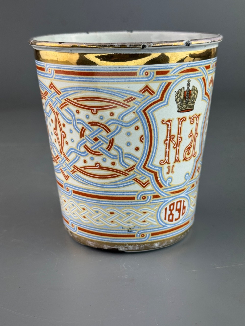 A white enamelled steel beaker decorated with red, blue, and gold geometric patterns, featuring the crowned cypher of Tsar Nicholas II and the date 1896. View V