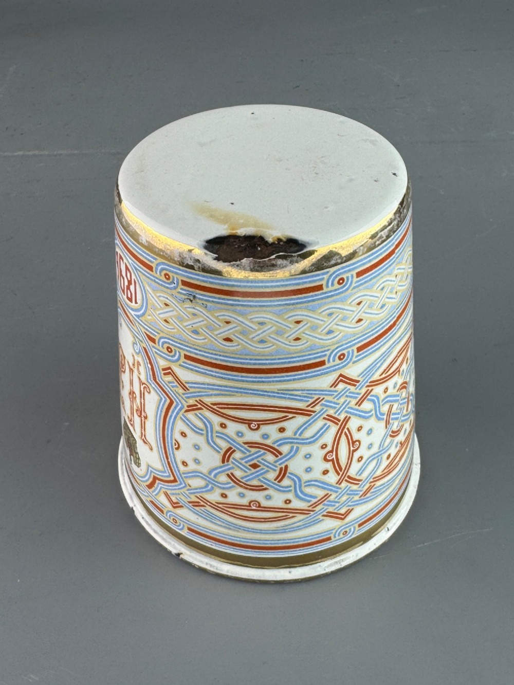 A white enamelled steel beaker decorated with red, blue, and gold geometric patterns, featuring the crowned cypher of Tsar Nicholas II and the date 1896. View VI