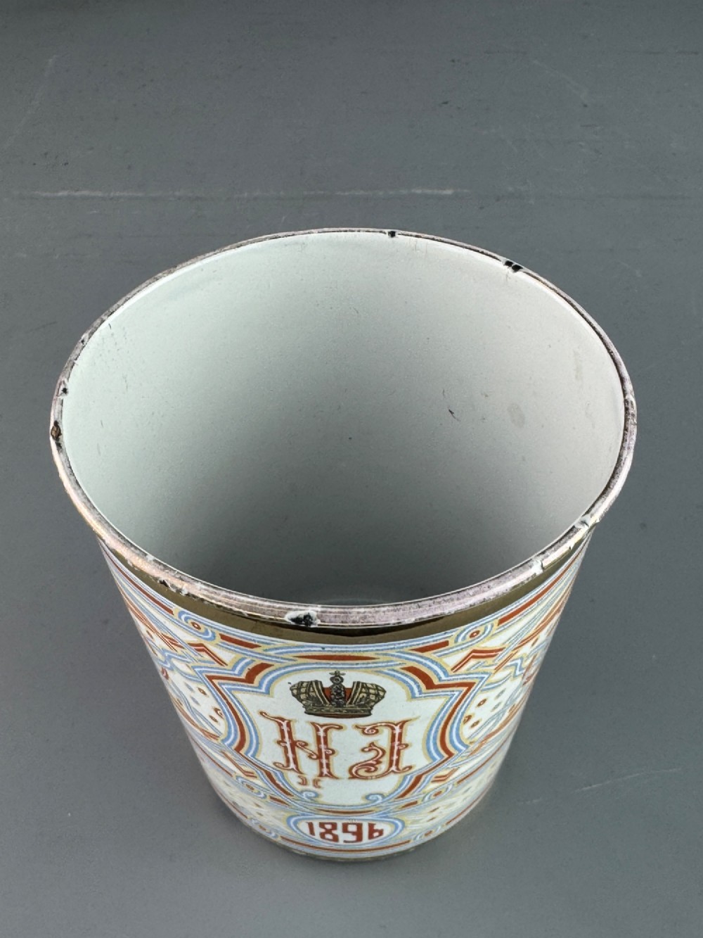 A white enamelled steel beaker decorated with red, blue, and gold geometric patterns, featuring the crowned cypher of Tsar Nicholas II and the date 1896. View VII